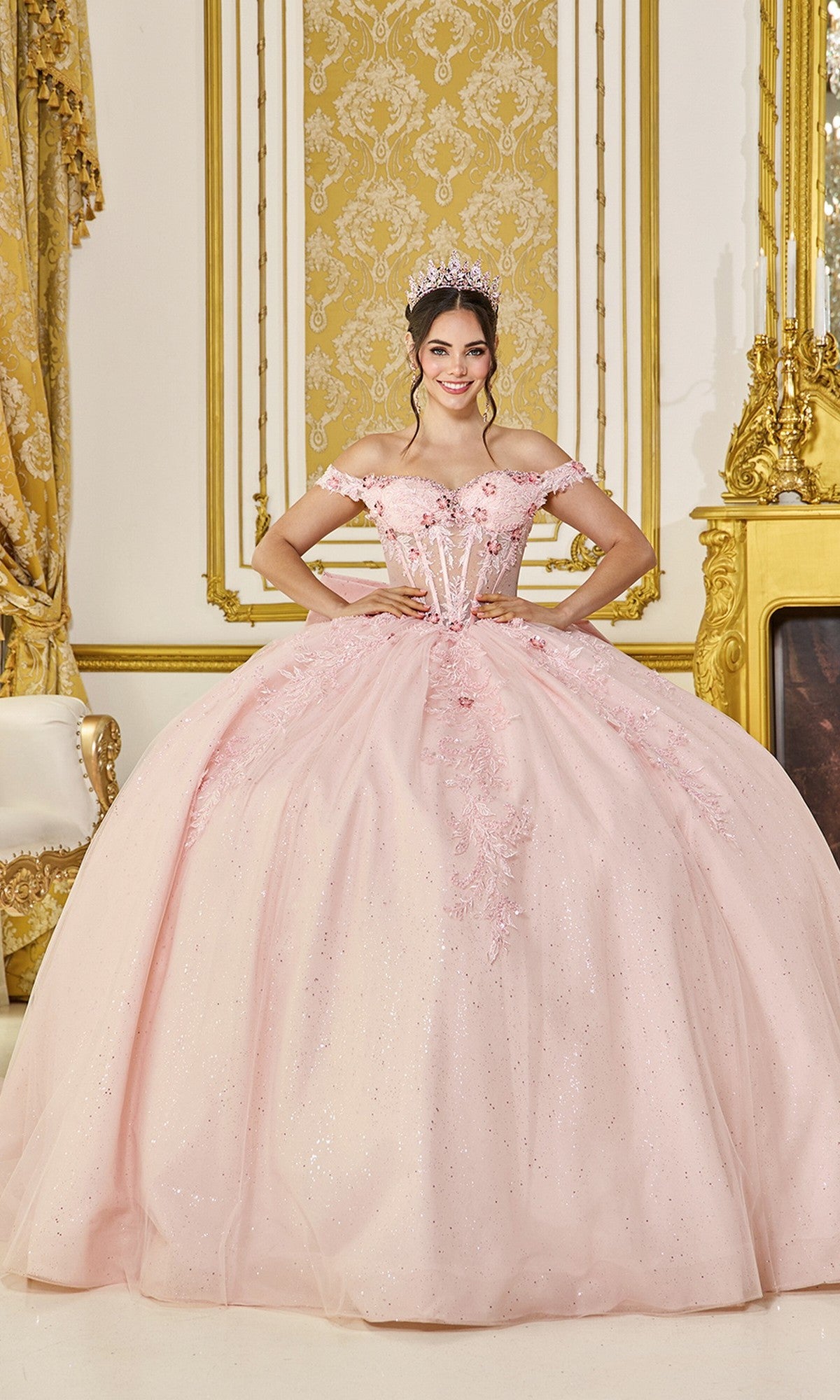 A young woman in the Quinceanera Dress 5090 by Dancing Queen, blush color, with glitter tulle and off-the-shoulder design, smiles in an ornate gold-decorated room while wearing a tiara.